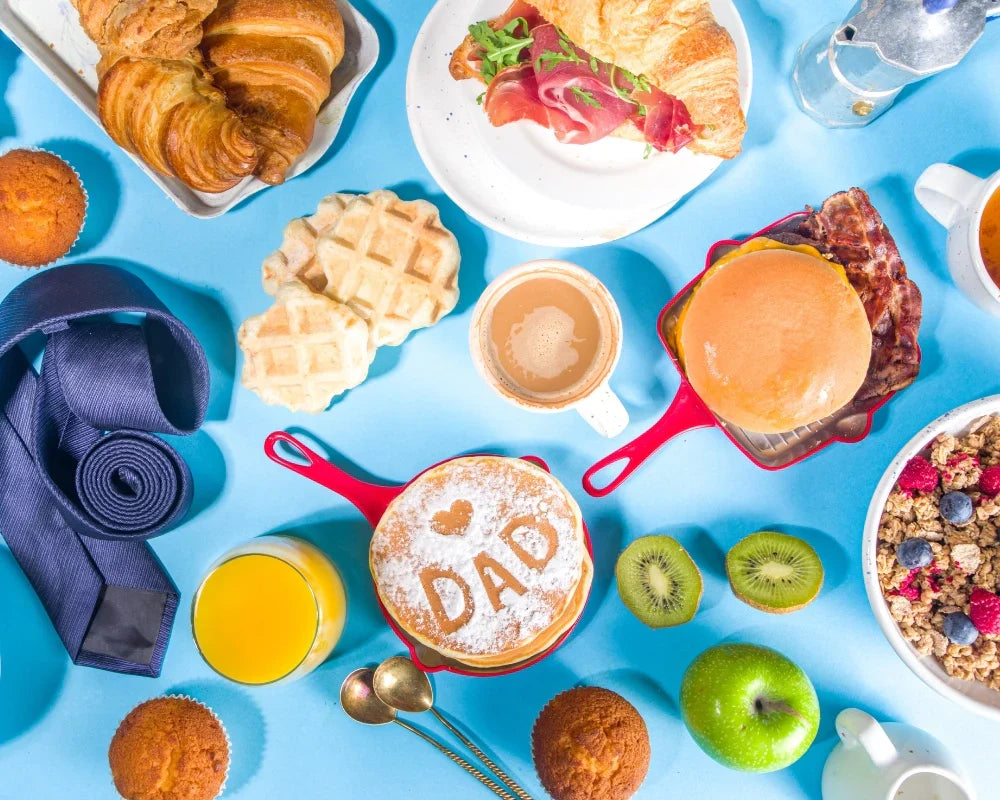 Colorful Father's Day breakfast setup with pancakes labeled 'DAD', croissants, coffee, fruits, muffins, and a necktie on a bright blue tablecloth.