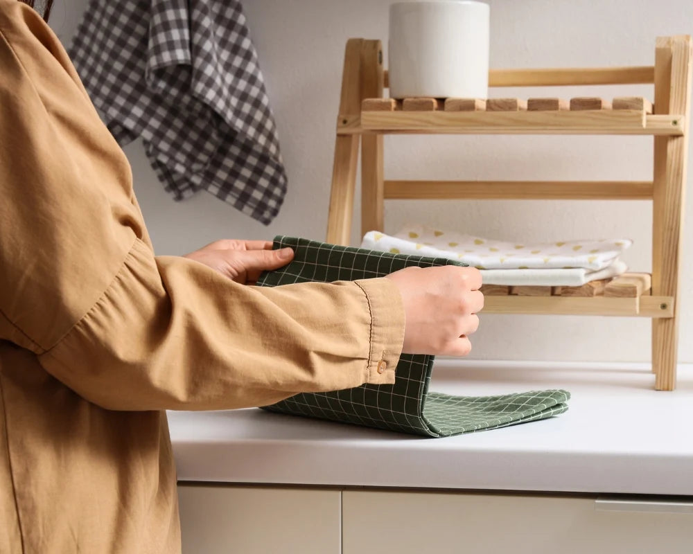 Person in a tan shirt folding a green checked dish towel beside a wooden rack holding more towels Person in a tan shirt folding a green checked dish towel beside a wooden rack holding more towels