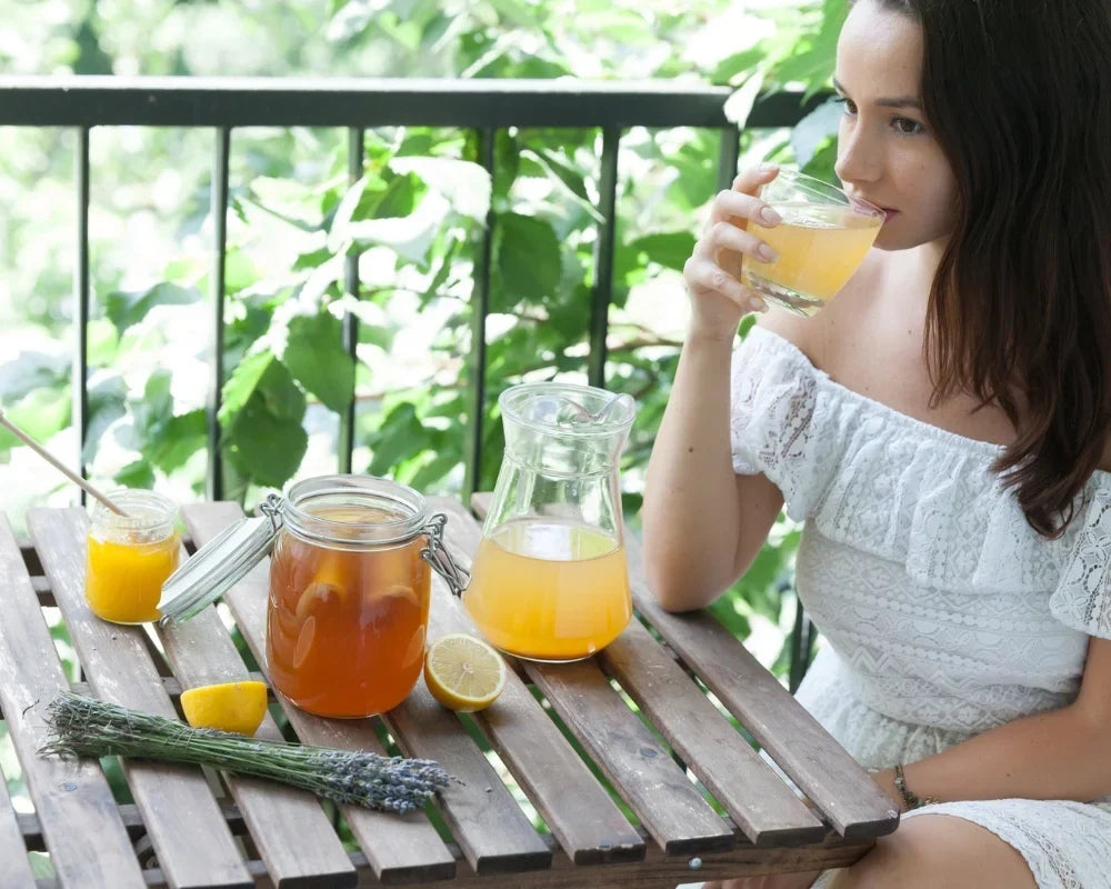 Woman sipping homemade kombucha at an outdoor wooden table with a glass jar and pitcher of kombucha Woman sipping homemade kombucha at an outdoor wooden table with a glass jar and pitcher of kombucha