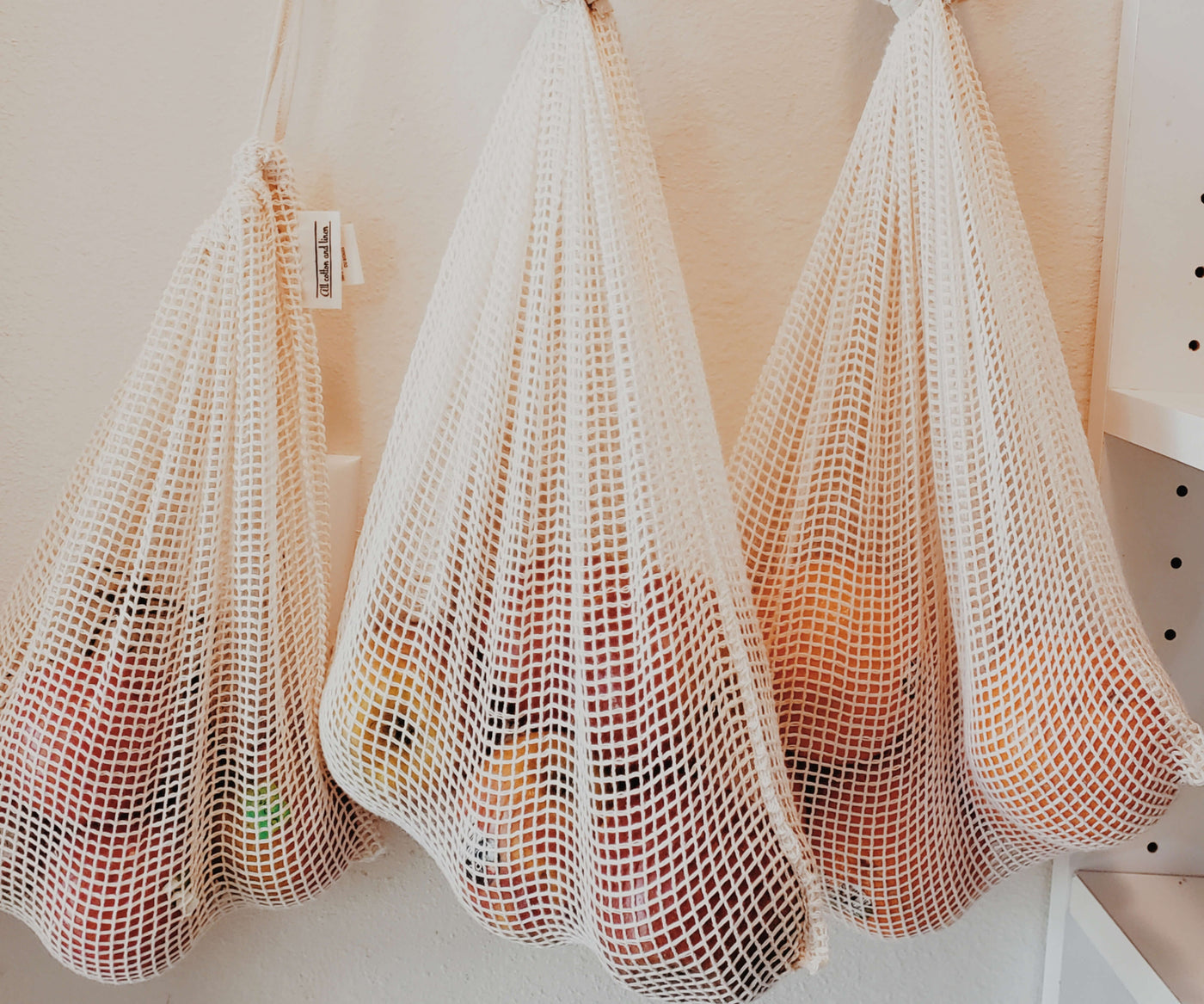 Three hanging white mesh bags filled with fruits like apples and oranges on a wall. Three hanging white mesh bags filled with fruits like apples and oranges on a wall.
