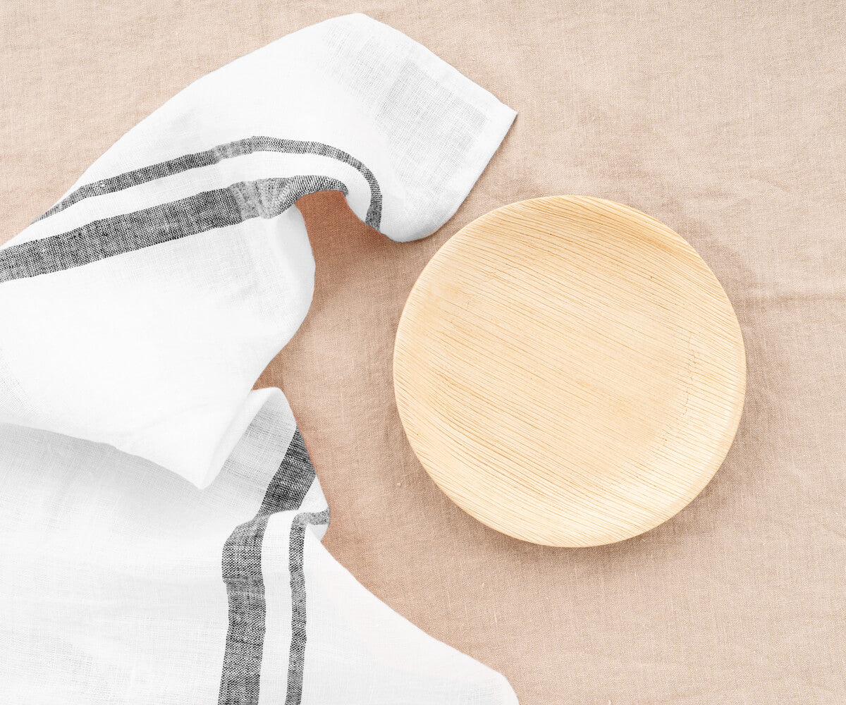 A black French linen tea towel is displayed next to a wooden plate on a linen surface, emphasizing its elegance and simplicity. A black French linen tea towel is displayed next to a wooden plate on a linen surface, emphasizing its elegance and simplicity.