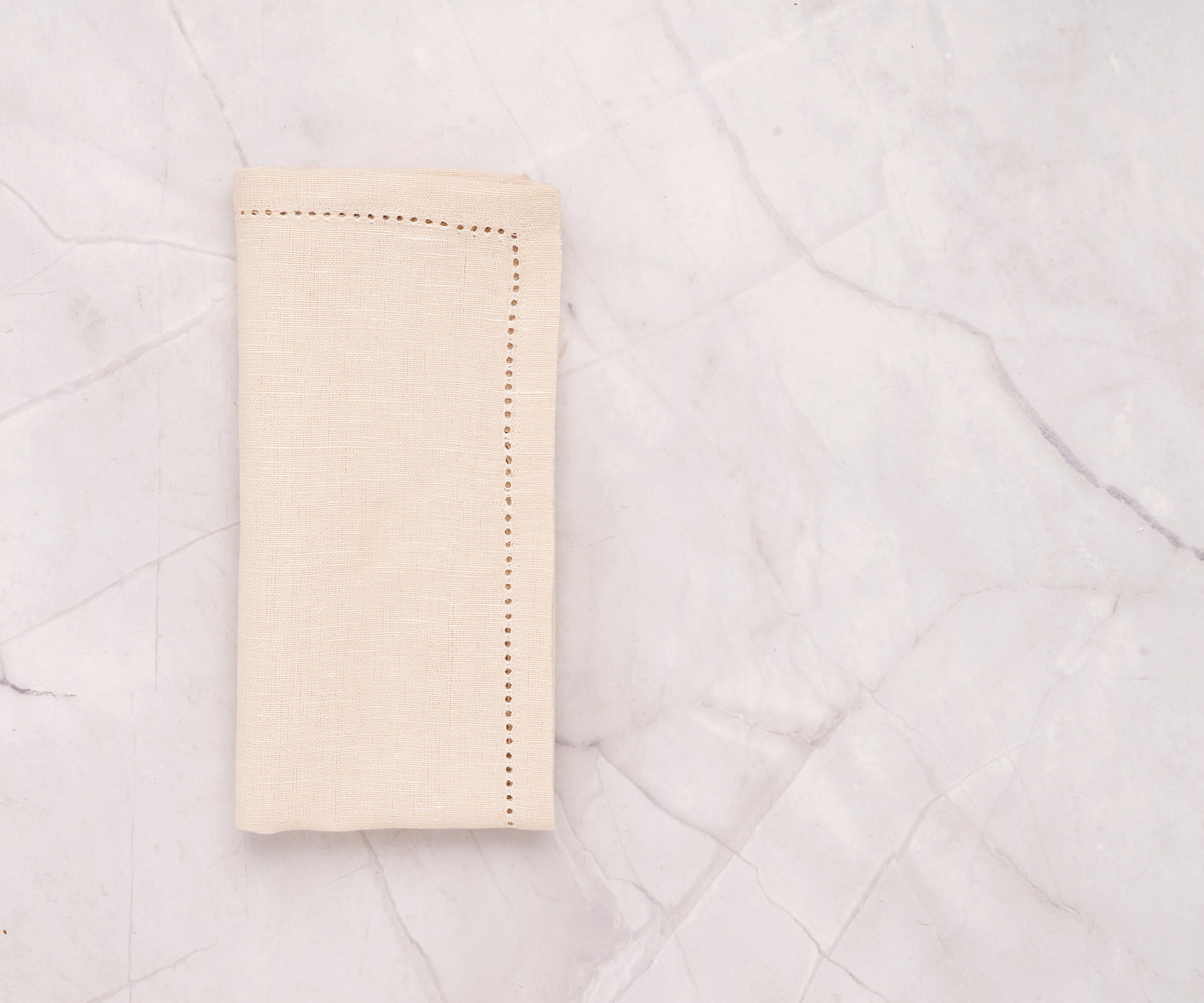 Natural hemstitch cotton linen napkins neatly folded on a dining table – soft beige napkins with elegant stitched border. Natural hemstitch cotton linen napkins neatly folded on a dining table – soft beige napkins with elegant stitched border.