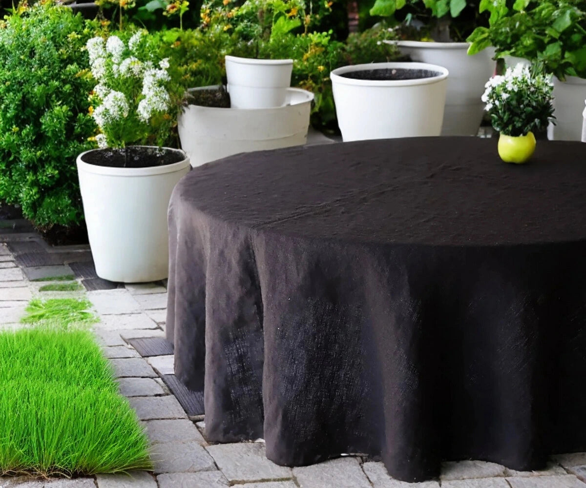 A black linen round tablecloth on a table outdoors, with chairs around it and plants in the background. A black linen round tablecloth on a table outdoors, with chairs around it and plants in the background.