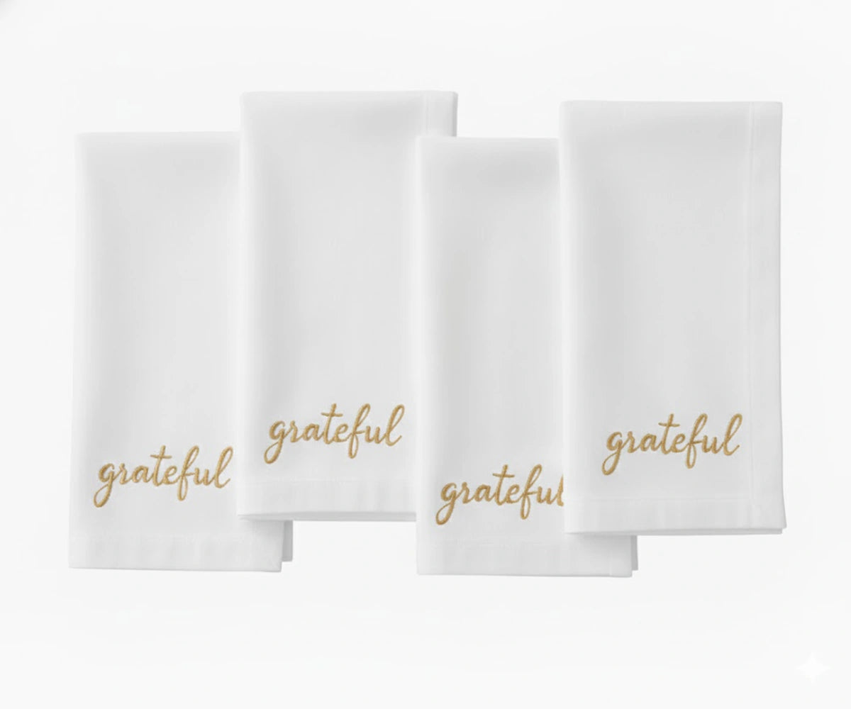 White cotton napkin with “Grateful” embroidered in beige thread for holiday table décor. White cotton napkin with “Grateful” embroidered in beige thread for holiday table décor.