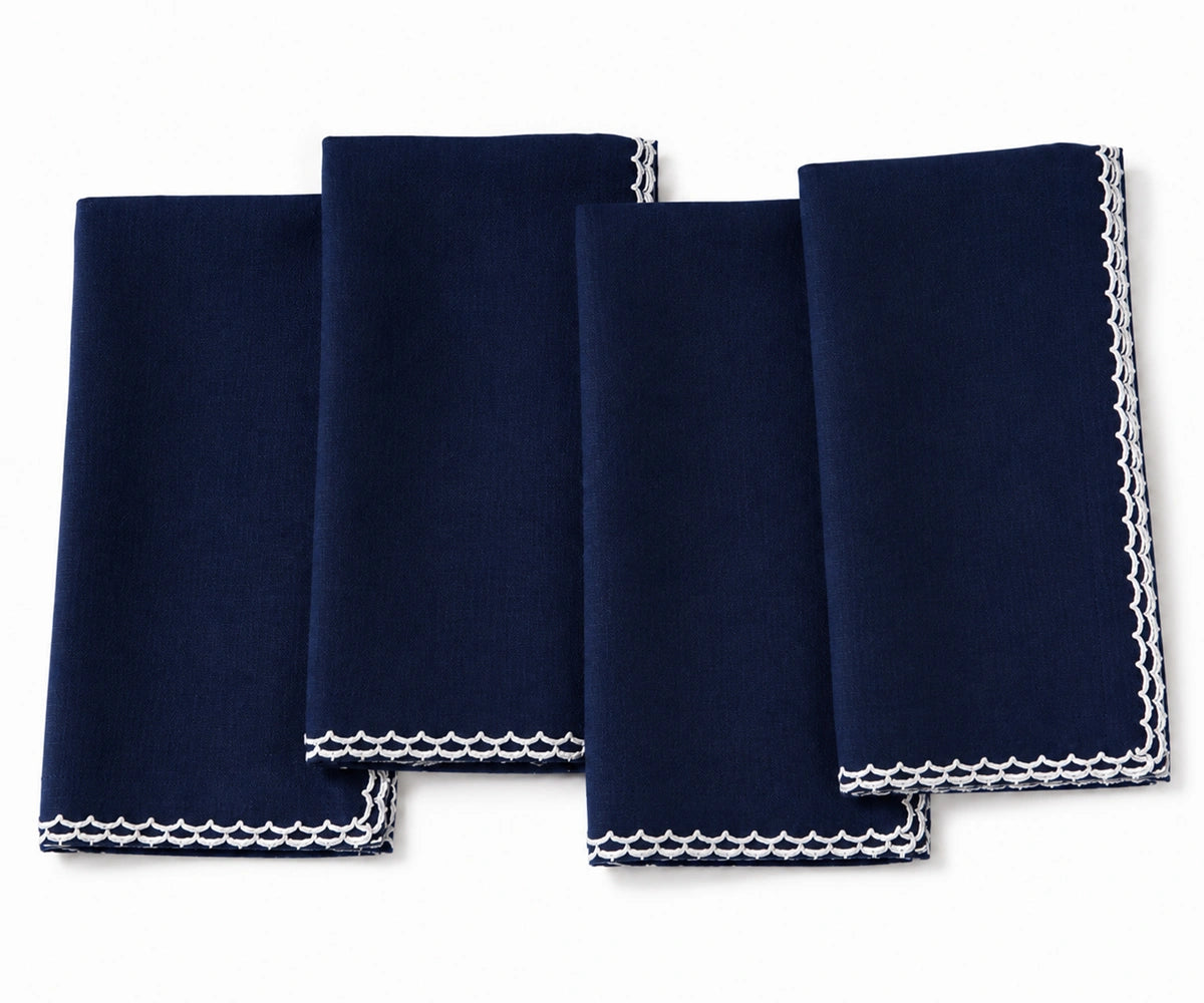Four navy blue napkins with white embroidered borders, arranged in an overlapping fan layout on a white background. Four navy blue napkins with white embroidered borders, arranged in an overlapping fan layout on a white background.
