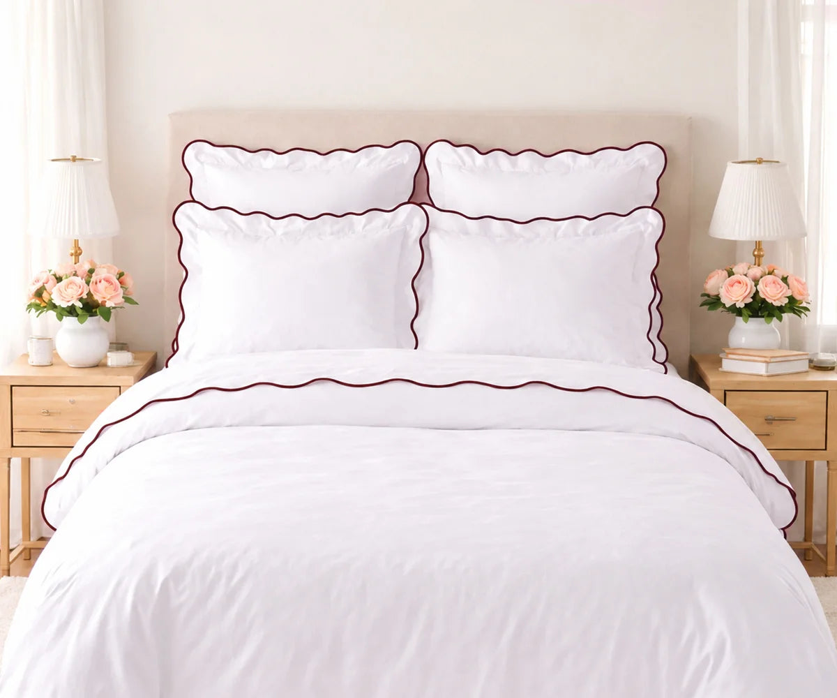White cotton bedding set with chocolate brown scalloped edge trim, including pillow shams and duvet cover, set on a bed flanked by wooden nightstands with lamps and pink flowers. White cotton bedding set with chocolate brown scalloped edge trim, including pillow shams and duvet cover, set on a bed flanked by wooden nightstands with lamps and pink flowers.