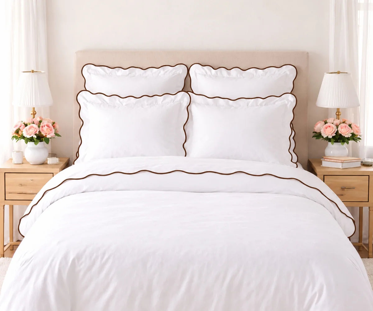 White cotton bedding set with coffee brown scalloped edge trim, including pillow shams and duvet cover, set on a bed flanked by wooden nightstands with lamps and pink flowers. White cotton bedding set with coffee brown scalloped edge trim, including pillow shams and duvet cover, set on a bed flanked by wooden nightstands with lamps and pink flowers.