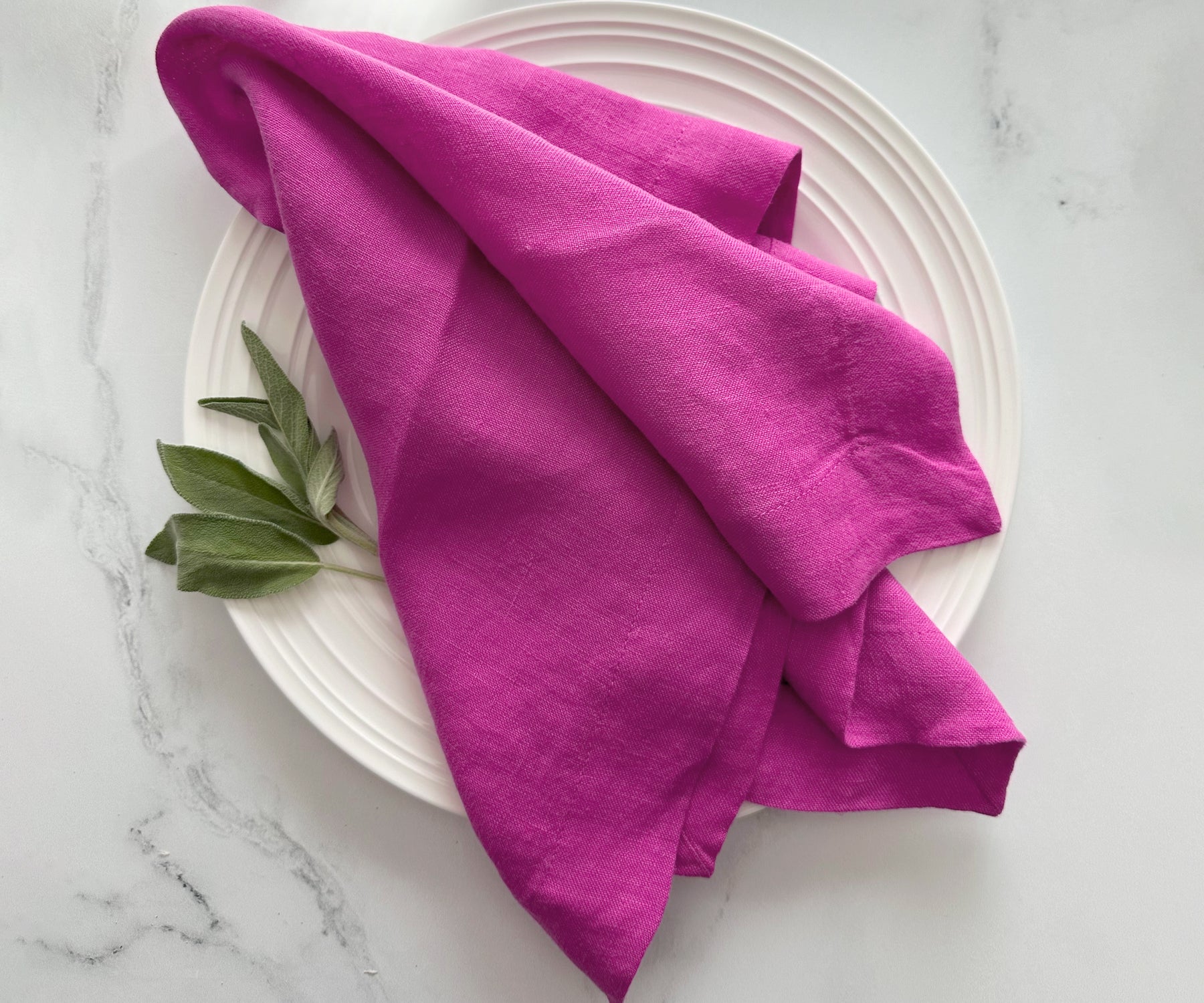 Orchid linen dinner napkin folded on a white plate with fresh sage leaves beside it. Orchid linen dinner napkin folded on a white plate with fresh sage leaves beside it.