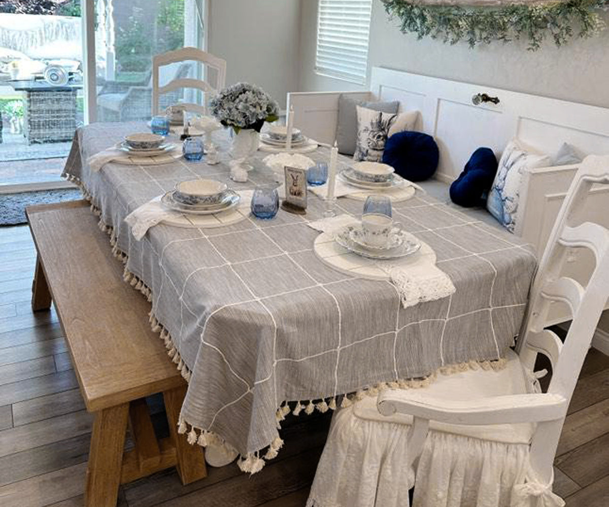 A rectangular cotton tablecloth with a plaid pattern and tassel details, displayed on a dining table with place settings and decorative chairs in the background. A rectangular cotton tablecloth with a plaid pattern and tassel details, displayed on a dining table with place settings and decorative chairs in the background.