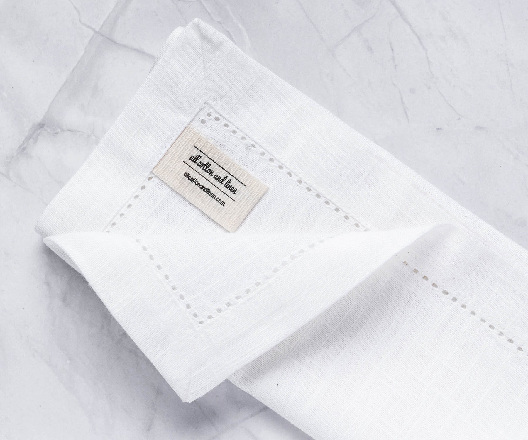 A folded white cotton napkin with borders with the label of All cotton and linen. A folded white cotton napkin with borders with the label of All cotton and linen.