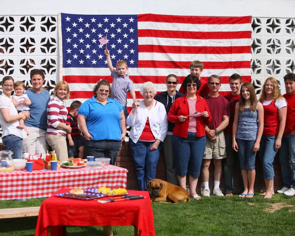 Large multigenerational family posing in front of an American flag backdrop during a 4th of July celebration with picnic tables and food in the foreground.