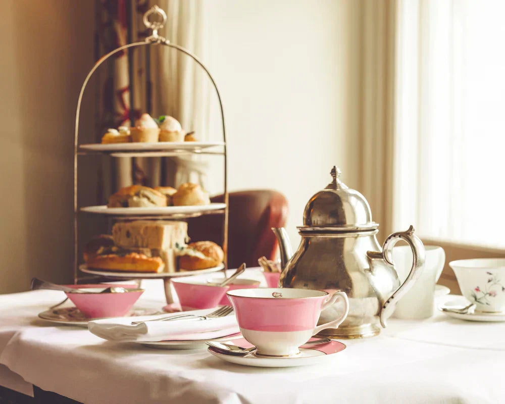 Tea set with pink teacup and saucer, silver teapot, and pastries on a table.
