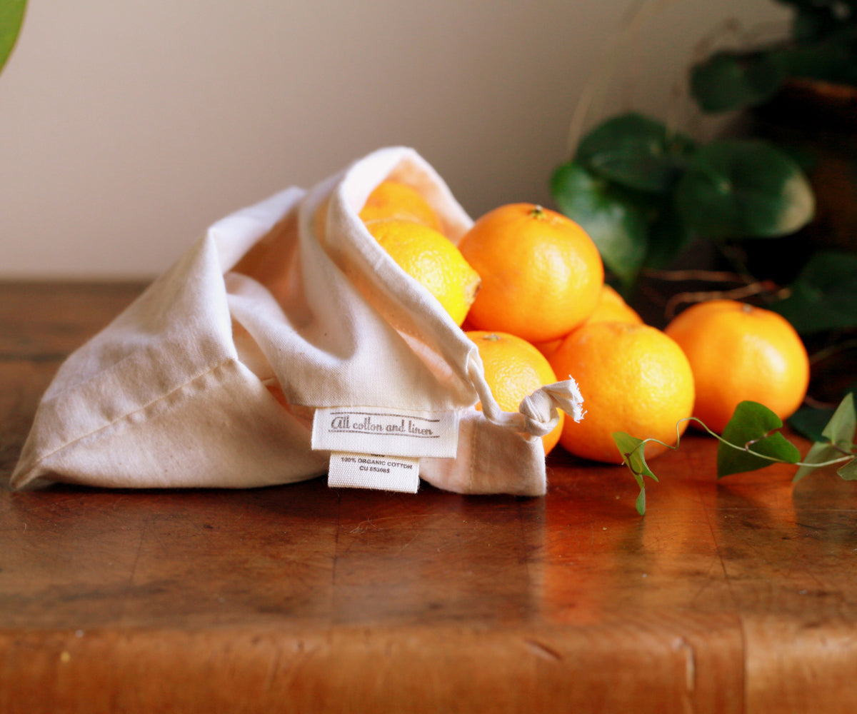 Various sizes and designs of muslin bags made from natural cotton fabric, displayed on a rustic wooden table with dried herbs, handmade soaps, and fresh produce.