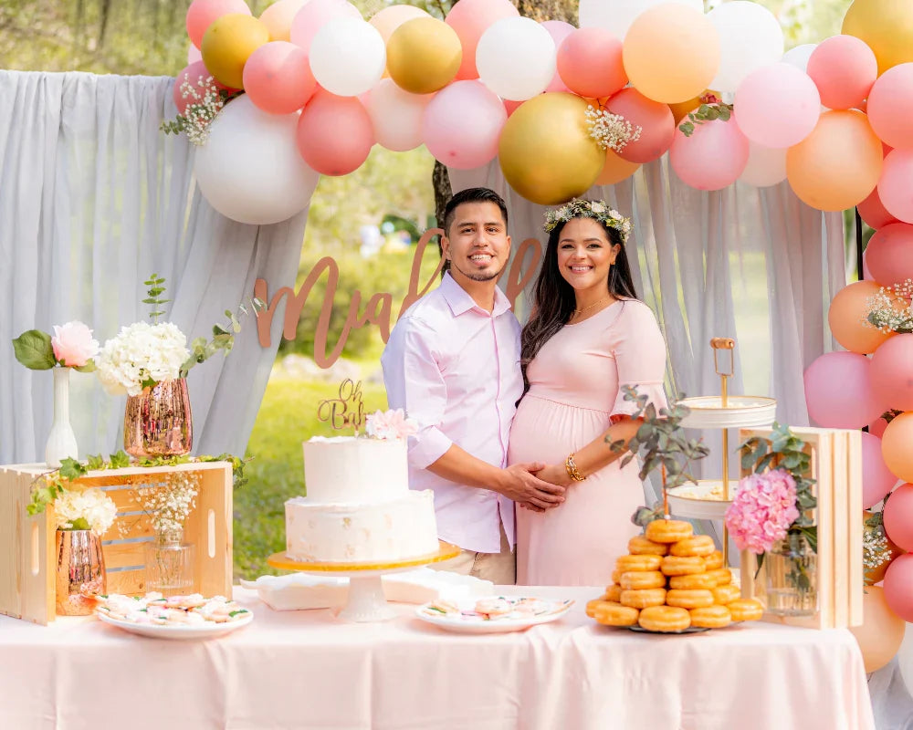 Couple at a baby shower with pink and gold decor, standing by a dessert table with cake, donuts, flowers, and a balloon arch.