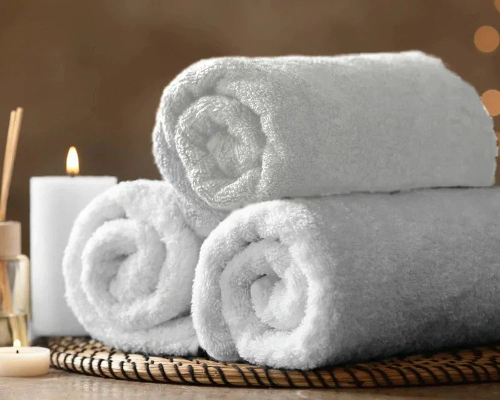 Soft white cotton bath towels rolled and stacked on a woven tray beside candles in a spa setting