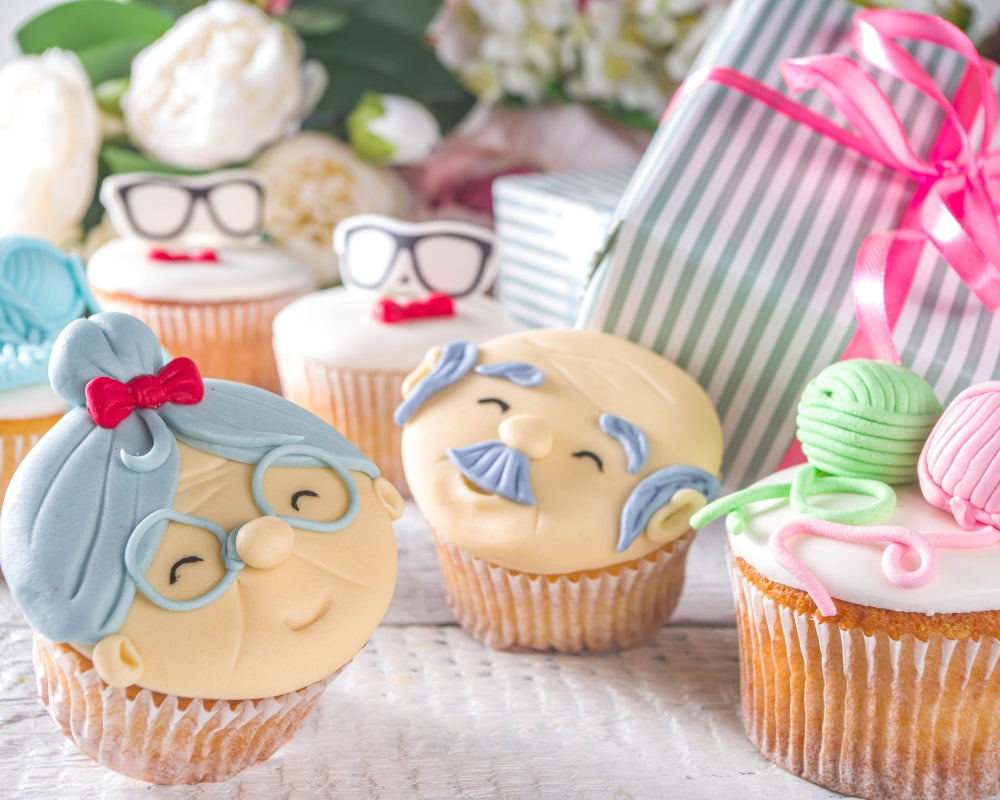 Colorful Grandparents Day cupcakes decorated with fondant faces of grandma and grandpa, yarn balls, and glasses, placed beside wrapped gifts and flowers — a sweet idea for celebrating and gifting grandparents.