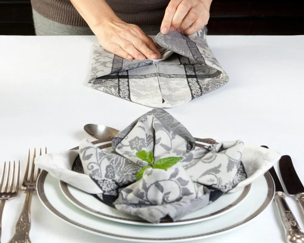 Person folding a gray and white checkered napkin over a bowl with silverware on a white table.