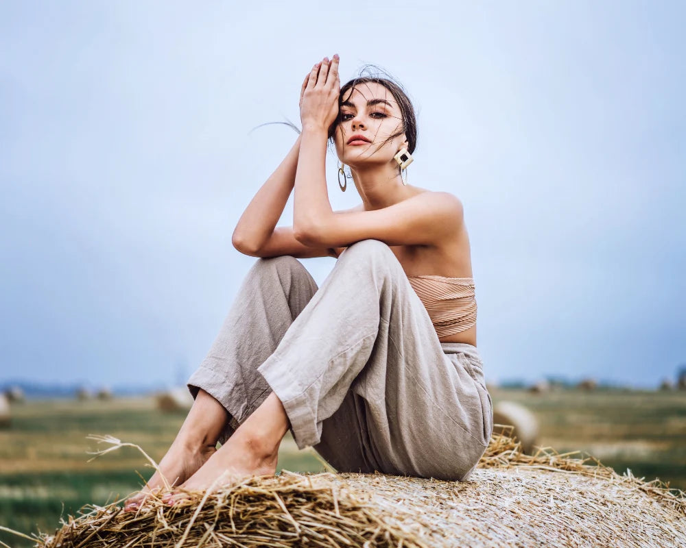 Woman wearing beige linen pants and a strapless top sitting barefoot on a hay bale in an open field, posing with one hand covering part of her face.