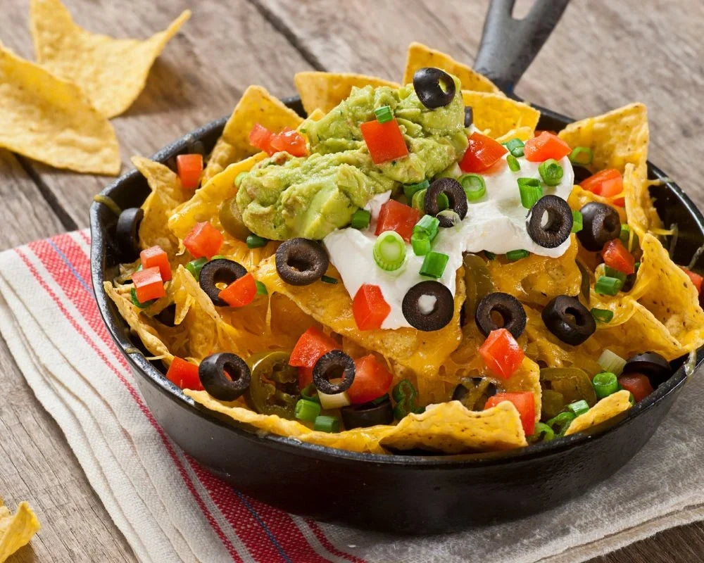 Cast iron skillet nachos topped with guacamole, sour cream, black olives, and tomatoes on a striped cloth