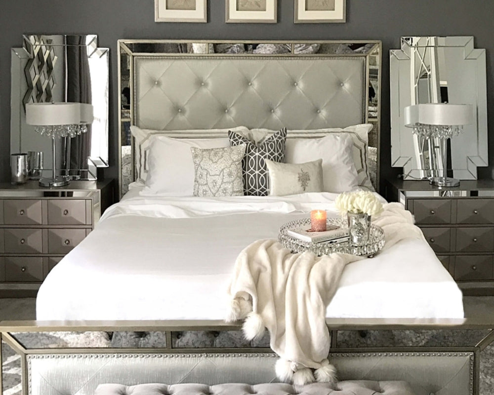 Elegant queen-size bed with white sheets, decorative pillows, mirrored nightstands, and a tufted headboard in a modern grey bedroom.