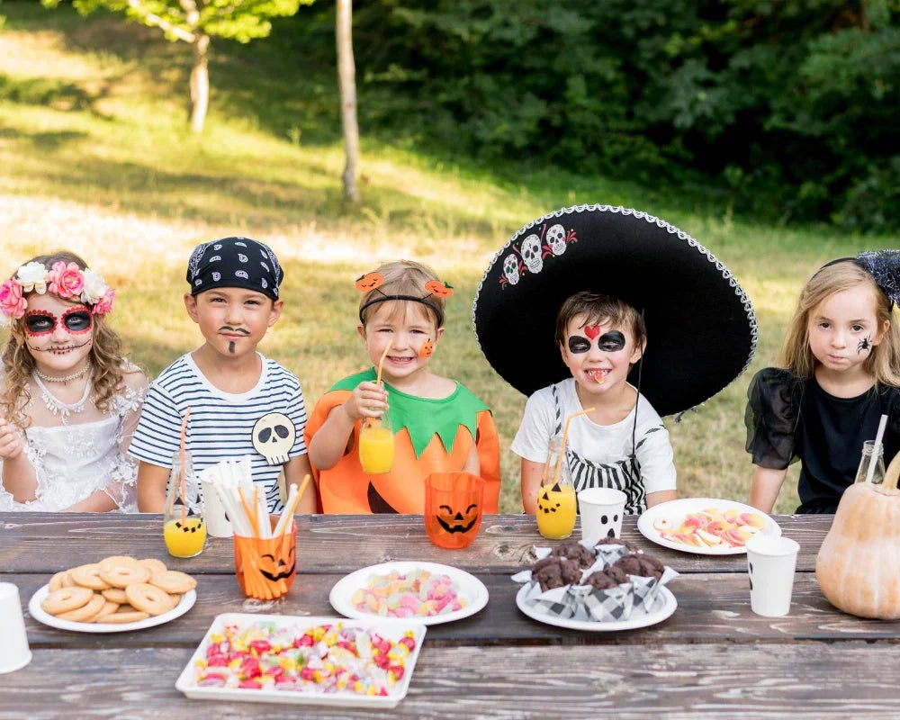 Group of kids in Halloween costumes enjoying snacks and drinks at an outdoor Summerween party table with themed treats and pumpkin cups.