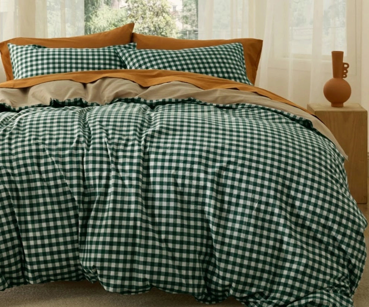 A cozy bedroom setting featuring a green and white gingham checkered duvet cover and matching pillows, layered with solid rust-orange pillowcases and a tan top sheet.