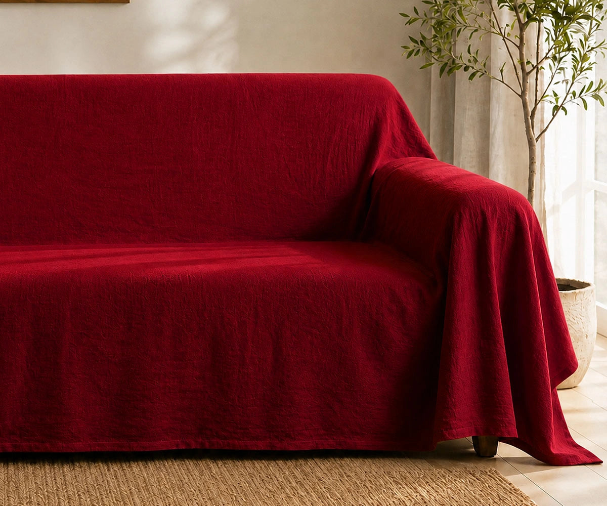 A deep red textured sofa slipcover draped over a couch in a bright, modern living room setting with a potted plant and light-colored walls.