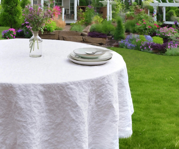 A white linen round tablecloth on a table outdoors, with chairs around it and plants in the background.
