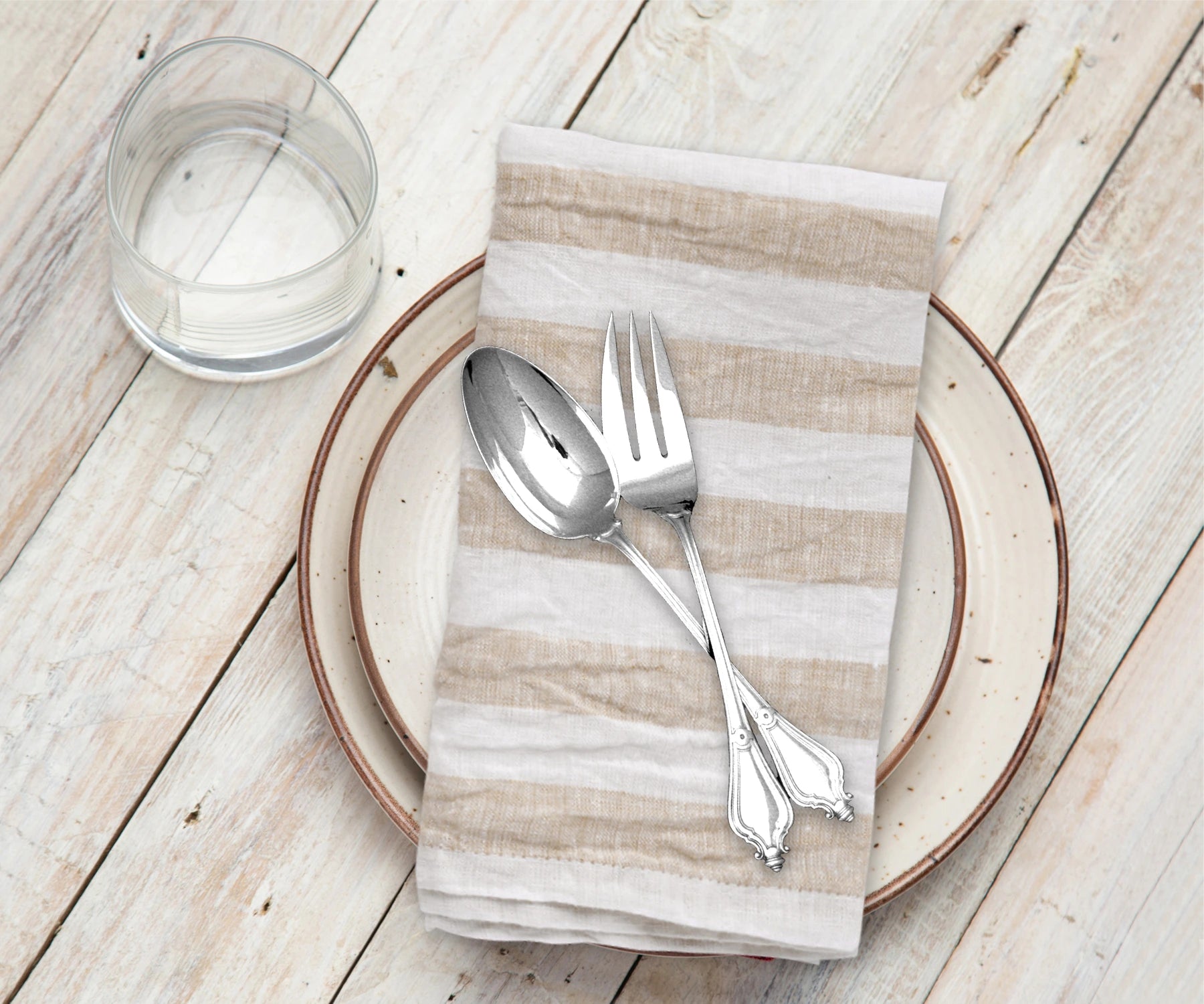 Silver spoon and fork on a striped napkin with a glass of water on a wooden table.