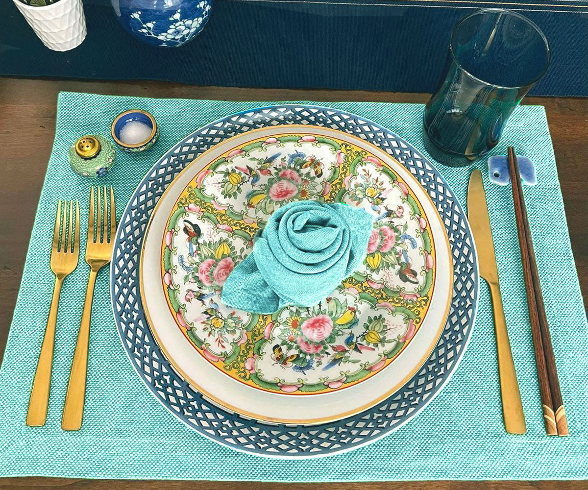 Elegant table setting with tonal napkins, floral patterned plates, turquoise placemat, gold cutlery, and chopsticks.