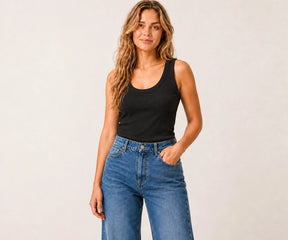 A woman modeling a black scoop-neck tank top paired with blue jeans, standing against a plain light gray studio background.