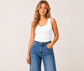 A woman modeling a white ribbed scoop-neck tank top paired with high-waisted blue jeans, standing against a plain light beige studio background.