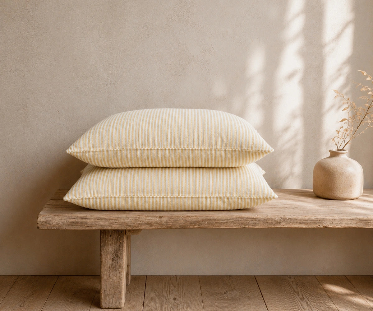 Beige striped linen pillowcases stacked on a rustic wooden bench with a ceramic vase in a warm, minimal bedroom.
