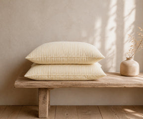 Beige striped linen pillowcases stacked on a rustic wooden bench with a ceramic vase in a warm, minimal bedroom.