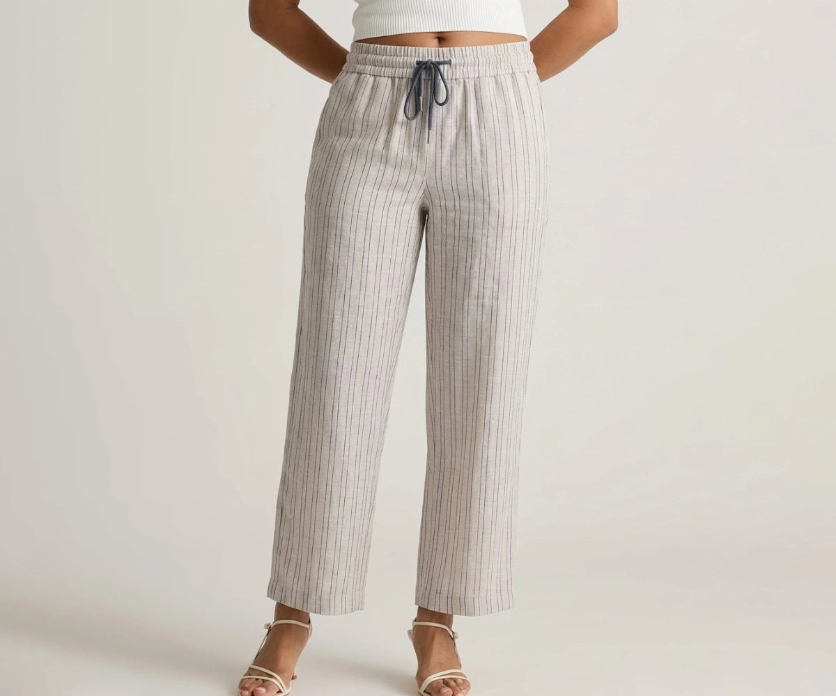 Front view of a model wearing high-waisted beige wide-leg cotton linen trousers with thin vertical pinstripes, featuring a dark blue drawstring and paired with white strappy sandals.