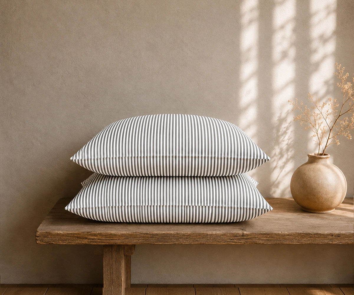 Black and white striped linen pillow covers stacked on a rustic wooden bench with a ceramic vase in a minimal bedroom setting.