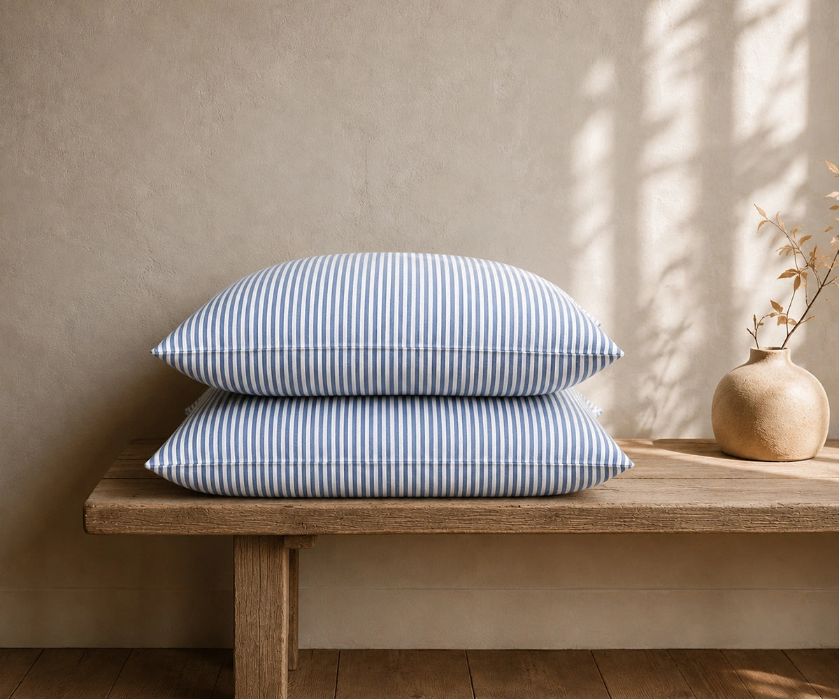  Blue and white striped linen pillowcases stacked on a rustic wooden bench with a ceramic vase in a minimal room.