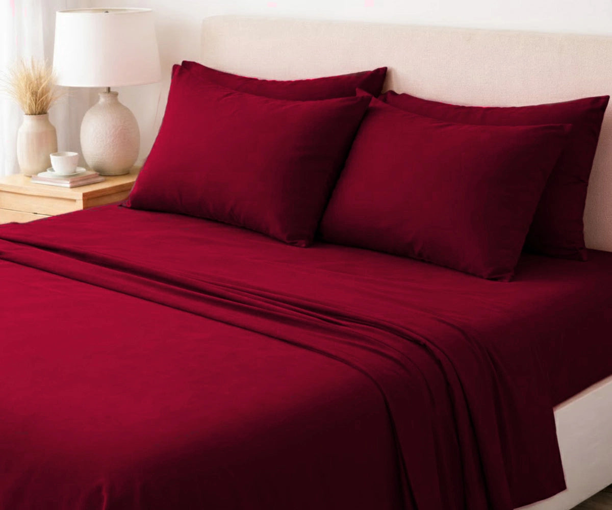 Burgundy bed sheets and pillows on a neatly made bed with a lamp and decor items on a nightstand.