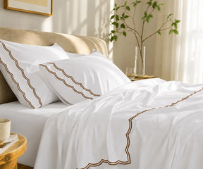 White scalloped bedding set with coffee brown trim styled on a bed in a sunlit bedroom