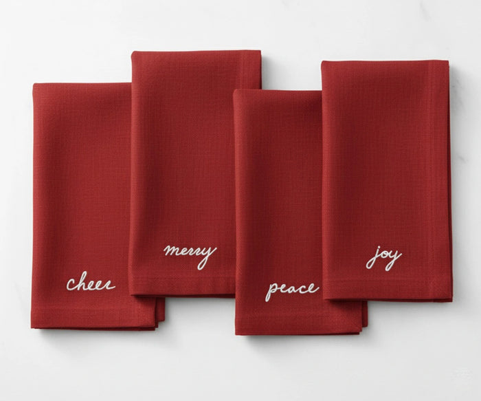 Red cotton napkin embroidered in white with festive Christmas words—Peace, Joy, Merry, and Cheer.