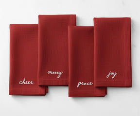 Red cotton napkin embroidered in white with festive Christmas words—Peace, Joy, Merry, and Cheer.