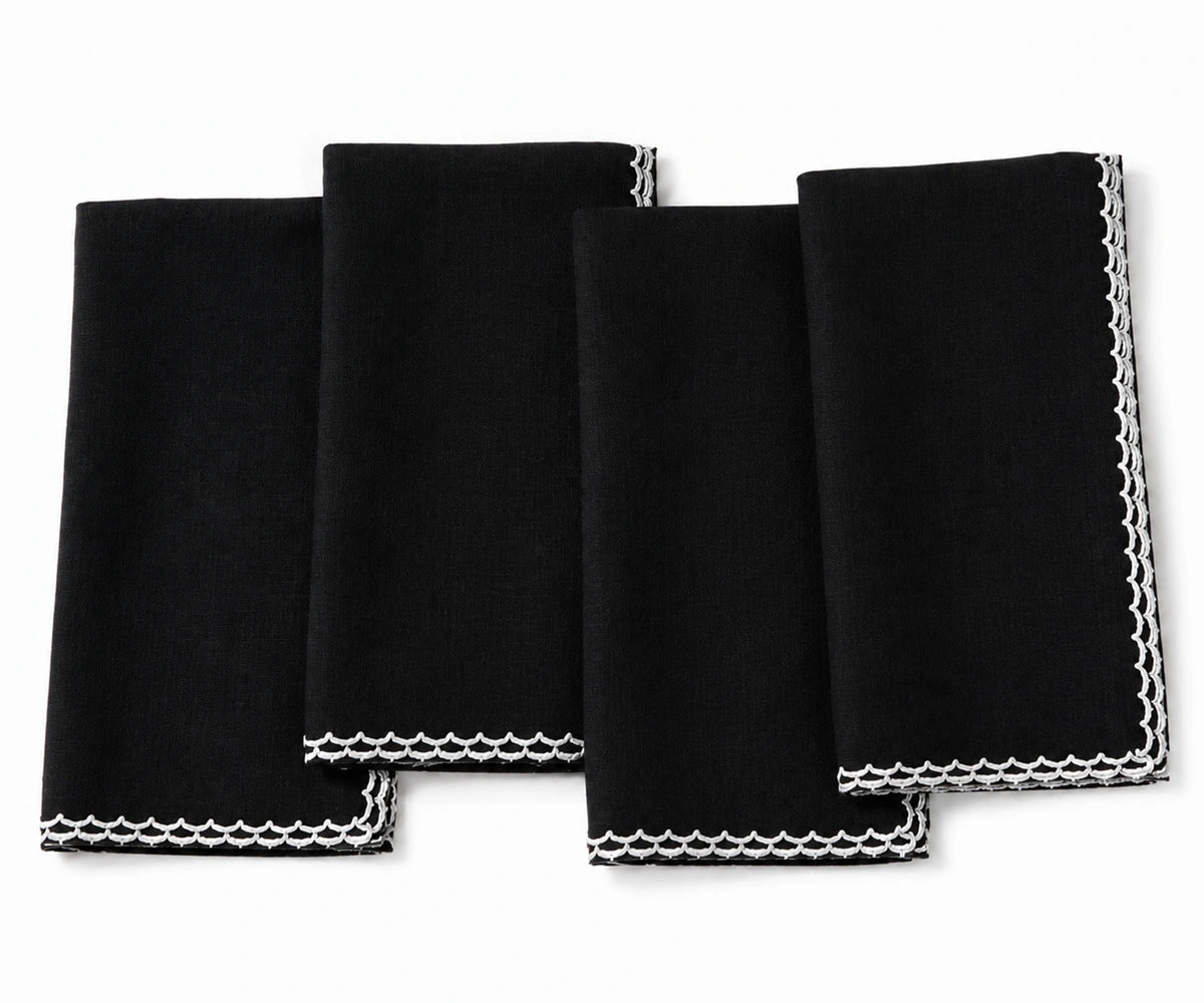 Four black square cloth napkins with white scalloped edges, arranged overlapping on a plain white background.