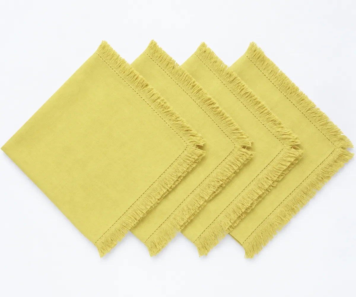 Butter yellow cotton napkins with decorative fringe trim styled beside white dinnerware and floral accents for a spring dining table.