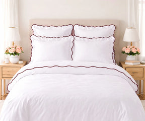 White cotton bedding set with chocolate brown scalloped edge trim, including pillow shams and duvet cover, set on a bed flanked by wooden nightstands with lamps and pink flowers.