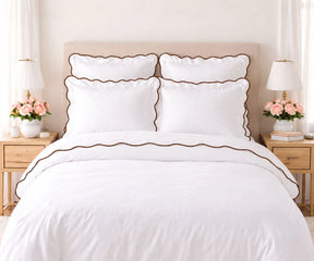 White cotton bedding set with coffee brown scalloped edge trim, including pillow shams and duvet cover, set on a bed flanked by wooden nightstands with lamps and pink flowers.