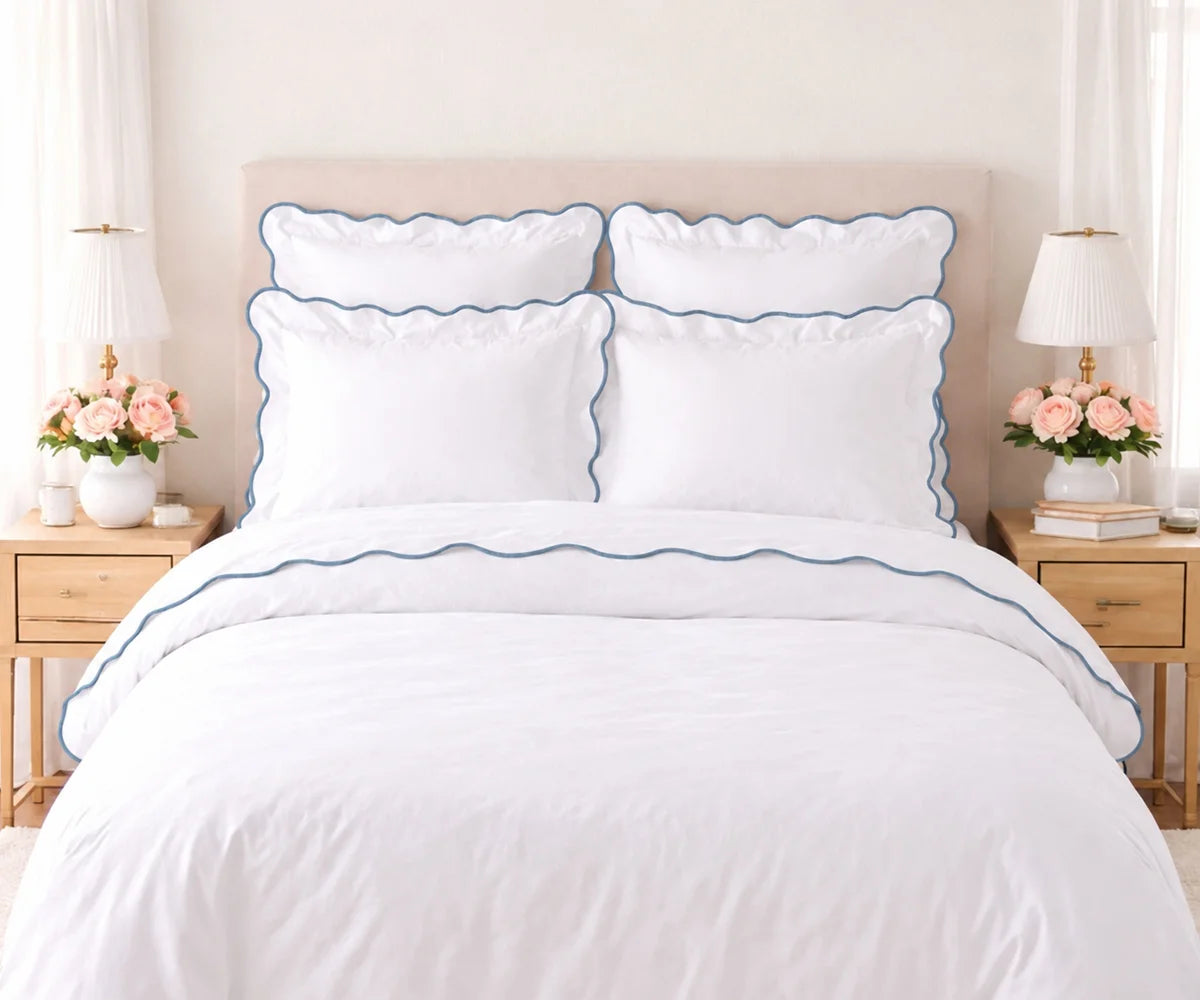 White cotton bedding set with dusty blue scalloped edge trim, including pillow shams and duvet cover, set on a bed flanked by wooden nightstands with lamps and pink flowers.