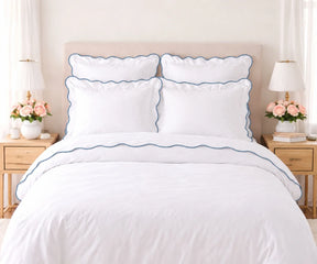 White cotton bedding set with dusty blue scalloped edge trim, including pillow shams and duvet cover, set on a bed flanked by wooden nightstands with lamps and pink flowers.