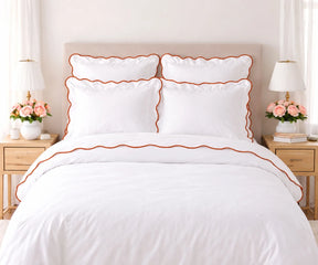 White cotton bedding set with terracotta orange scalloped edge trim, including pillows and duvet cover, set on a bed flanked by wooden nightstands with lamps and pink flower arrangements.