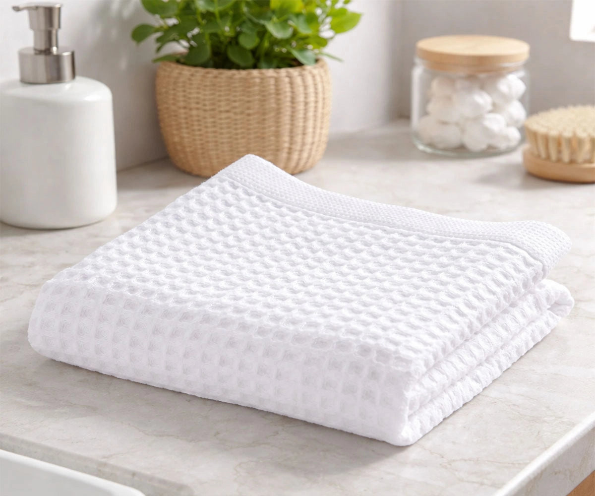 A neatly folded white waffle weave cotton towel on a marble bathroom countertop next to a soap dispenser and potted plant.