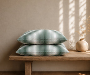  Forest green and white striped linen pillowcases stacked on a rustic wooden bench with a ceramic vase in a warm, minimal bedroom.